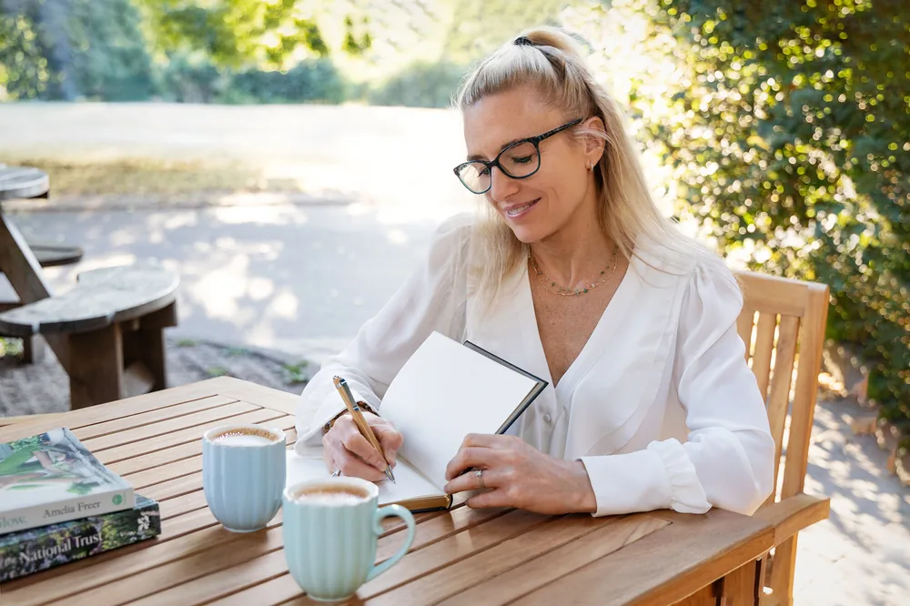 Charlotte Sinclair writing in a notebook at a garden table, two cups of coffee beside her.