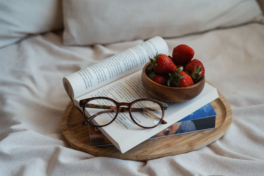 An open book with reading glasses laid across it, on a wooden tray with a small bowl of strawberries.
