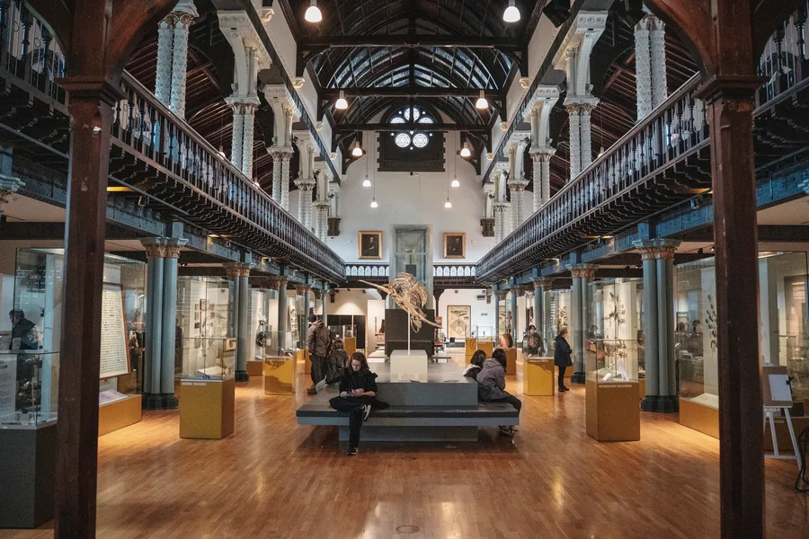 A tall galleried museum hall with a wooden floor and daylight from above, a few people sitting quietly on a bench in the middle.